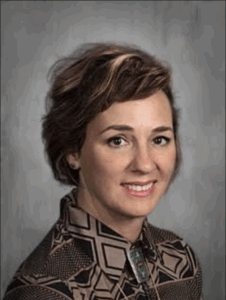 Woman with short, styled hair, wearing a patterned blouse, smiles against a gray background.