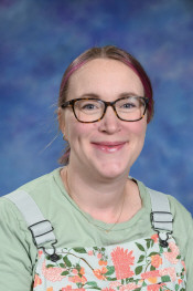 Smiling person with glasses and pink hair, wearing a floral shirt and overalls, against a blue background.