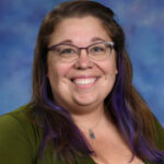 Smiling woman with glasses and purple-tinted hair, wearing a green cardigan, against a blue background.