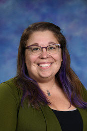 Smiling woman with glasses and purple-tinted hair, wearing a green cardigan, against a blue background.