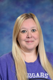 A woman with long blonde hair, wearing a purple shirt, smiles in front of a blue and purple backdrop.