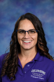 Smiling woman with long dark hair wearing glasses and a purple shirt, with a blurred blue background.