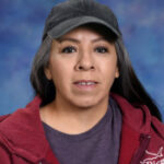 A woman with long dark hair wearing a black cap and a red hoodie, smiling against a blue background.