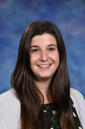 Smiling woman with long brown hair wearing a green dress and white cardigan, set against a blue background.