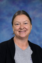 Smiling woman with shoulder-length hair, wearing a gray shirt and black cardigan, against a blurred blue background.