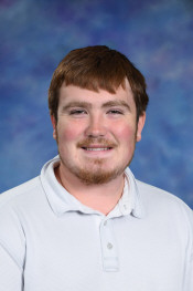 A young man with brown hair and a beard smiles for a photo against a blue and purple background.