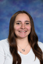 Smiling young woman with long, wavy brown hair, wearing a white top, against a blue and purple background.