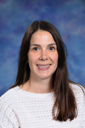 A woman with long brown hair smiles, wearing a white sweater, against a colorful blue background.