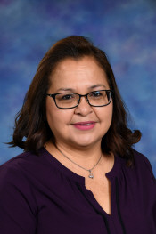 A woman with shoulder-length dark hair and glasses smiles, wearing a purple top, against a blue background.