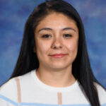 A woman with long black hair smiles, wearing a white sweater with blue stripes, against a blurred blue background.