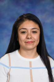 A woman with long black hair smiles, wearing a white sweater with blue stripes, against a blurred blue background.