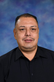 A man with short hair and a mustache, wearing a black shirt, poses against a blue background.