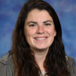 Smiling woman with long brown hair, wearing a gray jacket, against a blue and purple background.