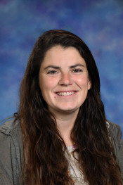 Smiling woman with long brown hair, wearing a gray jacket, against a blue and purple background.