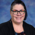 Smiling woman with short dark hair and colorful glasses, wearing a black cardigan over a patterned top, against a blue background.