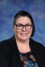 Smiling woman with short dark hair and colorful glasses, wearing a black cardigan over a patterned top, against a blue background.