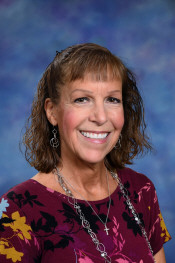 A smiling woman with curly hair, wearing a floral top and necklace, poses against a blue background.