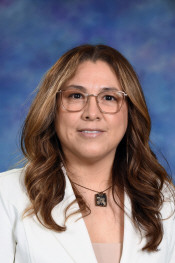 Portrait of a woman with long wavy hair, wearing glasses and a white blazer, against a blue background.
