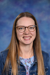 Smiling woman with long brown hair and glasses, wearing a denim jacket over a floral top, against a blue background.