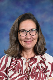 A woman with long brown hair and glasses smiles, wearing a patterned blouse against a blue background.