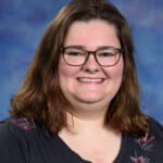 A smiling young woman with shoulder-length brown hair and glasses, wearing a dark shirt with floral details, against a blue background.