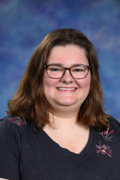 A smiling young woman with shoulder-length brown hair and glasses, wearing a dark shirt with floral details, against a blue background.