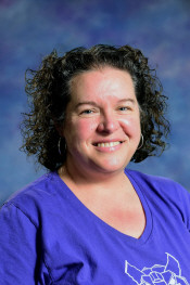 Smiling woman with curly hair wearing a purple shirt, against a blurred blue and purple background.