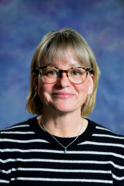 Smiling woman with short blonde hair, wearing glasses and a striped black-and-white shirt, against a colorful background.