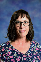 Woman with shoulder-length dark hair and glasses, wearing a floral-patterned shirt, against a blue background.
