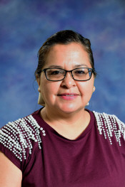 A woman with glasses and dark hair smiles, wearing a maroon shirt with decorative embellishments.