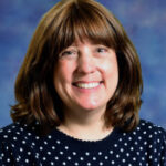 Una mujer sonriente con cabello castaño que lleva un suéter de lunares, sobre un fondo azul y morado.