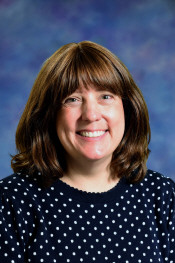 A smiling woman with brown hair wearing a polka dot sweater, set against a blue and purple background.