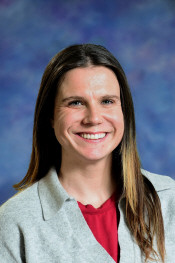 Una mujer sonriente con el pelo largo y castaño, vestida con un jersey gris sobre una camiseta roja, sobre un fondo azul.
