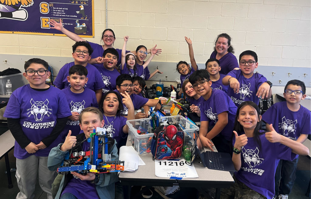 A group of smiling students in purple shirts pose with robotics projects at a table, showing excitement and teamwork.