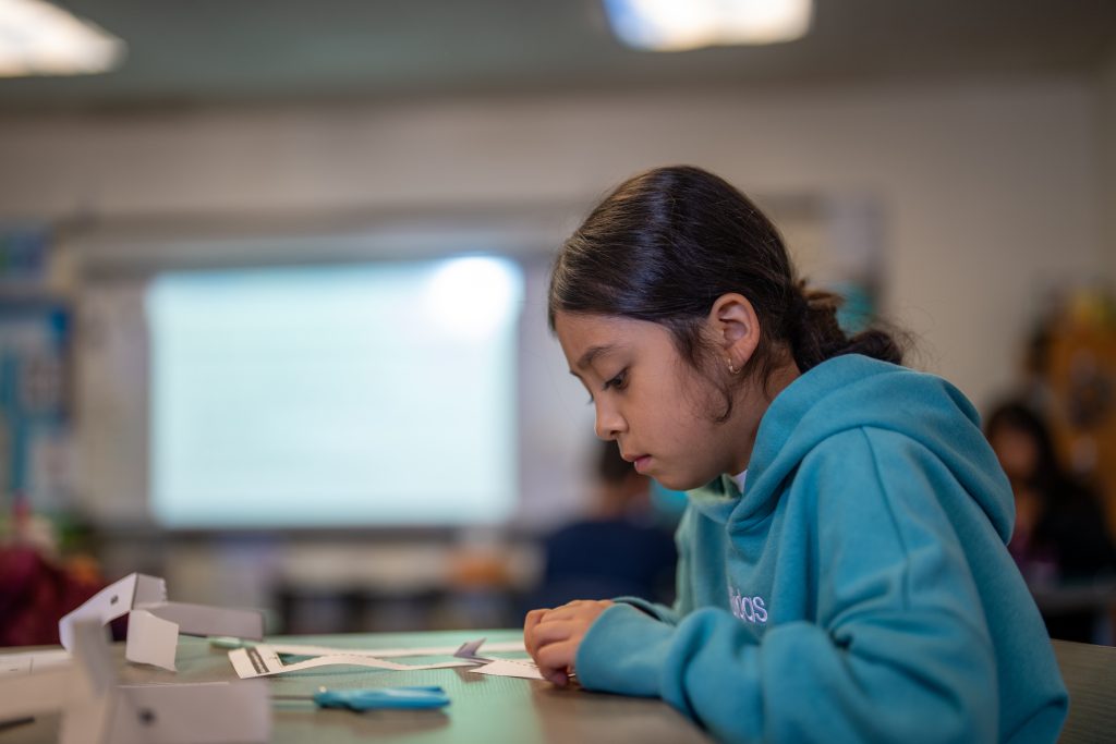 A girl in a teal hoodie focuses on paper cutouts at a classroom table, with a projector screen in the background.