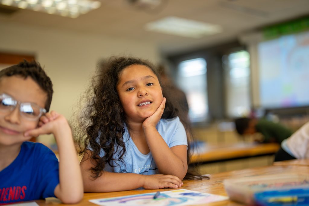 A smiling girl with curly hair rests her chin on her hand at a classroom table, with a boy in glasses beside her.