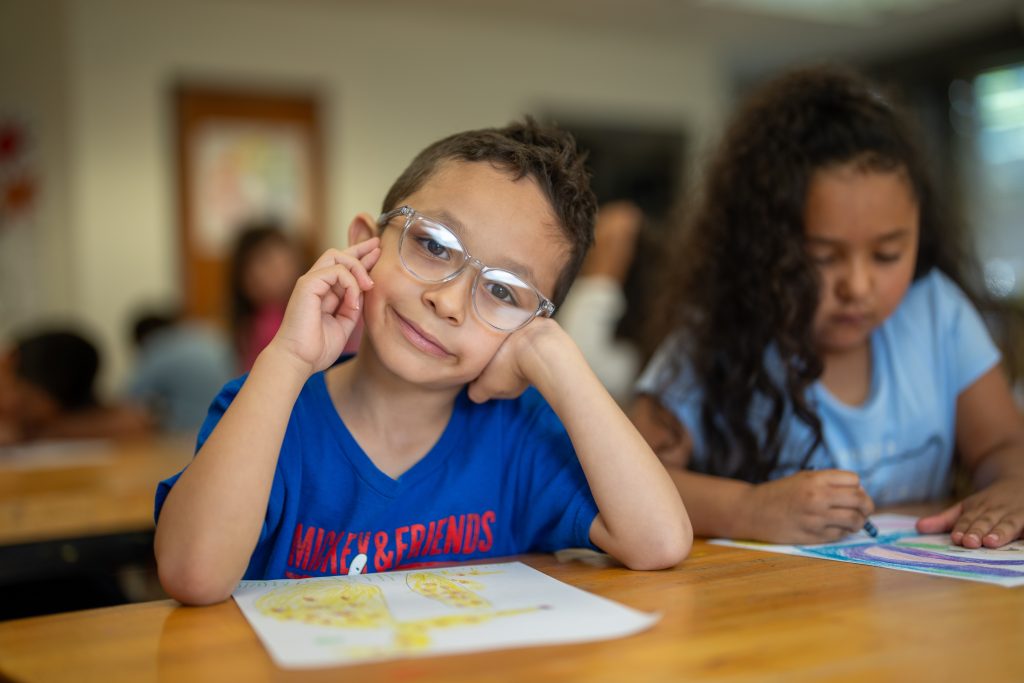 A young boy with glasses rests his chin on his hand, smiling while coloring at a classroom table with peers.