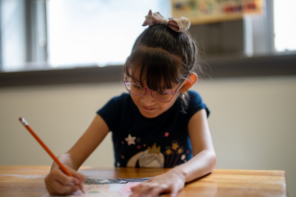 A young girl with glasses and a bow in her hair is drawing with a pencil at a wooden table.