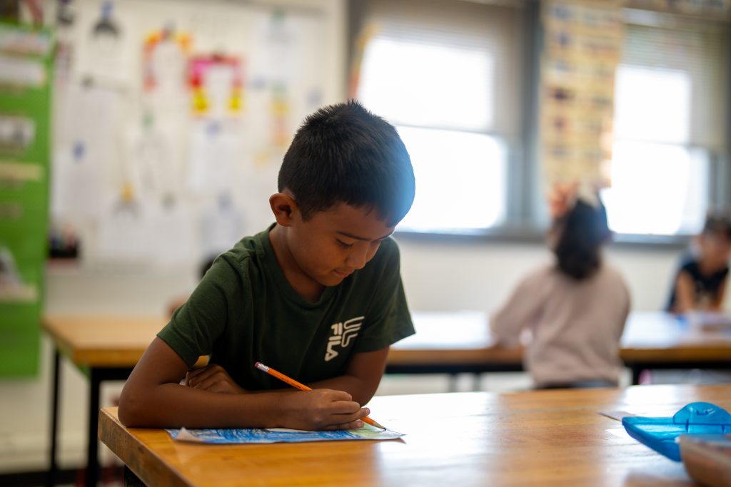 A young boy sits at a desk, focused on drawing with an orange pencil in a classroom setting.