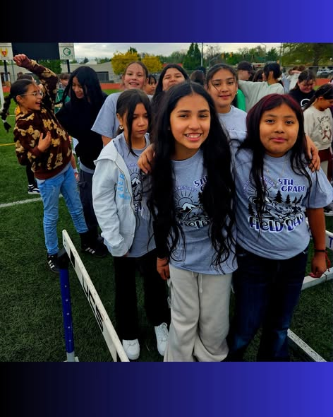 Un grupo de alumnos de quinto de primaria, todos sonrientes y con camisetas grises, está en un campo participando en un acto escolar.
