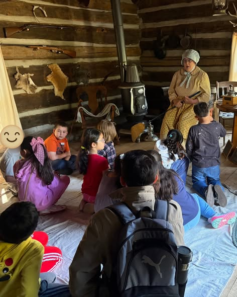 A group of children sits on the floor listening to an elderly woman in a rustic log cabin.