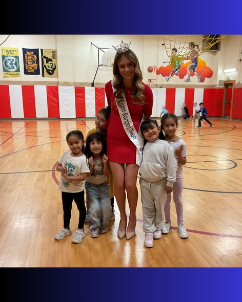 A crowned woman in a red dress poses with five smiling children in a gymnasium, colorful banners in the background.