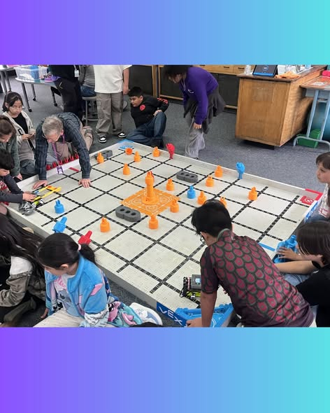 A group of children and adults gather around a large game board with orange and blue pieces, engaged in play.