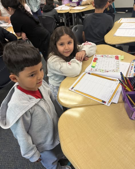 Two children stand at a classroom table with worksheets and colored pencils, engaged in an activity together.