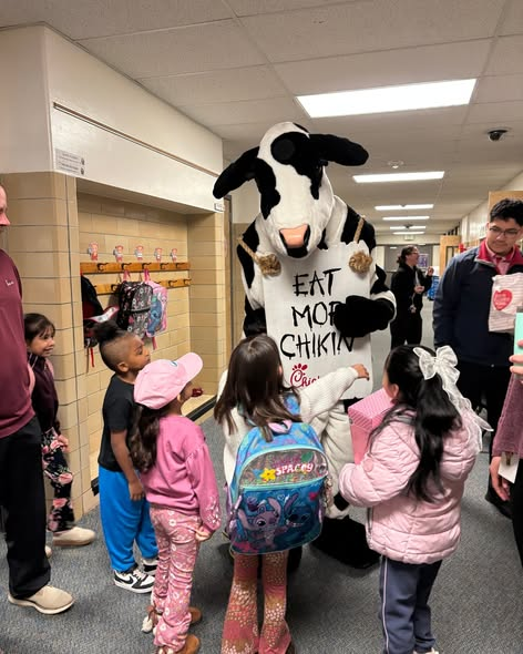 A costumed cow mascot stands in a school hallway surrounded by children, promoting a Chick-fil-A message.