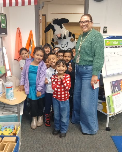 A group of children and a teacher smile for the camera, with a cow mascot in the background promoting healthy eating.