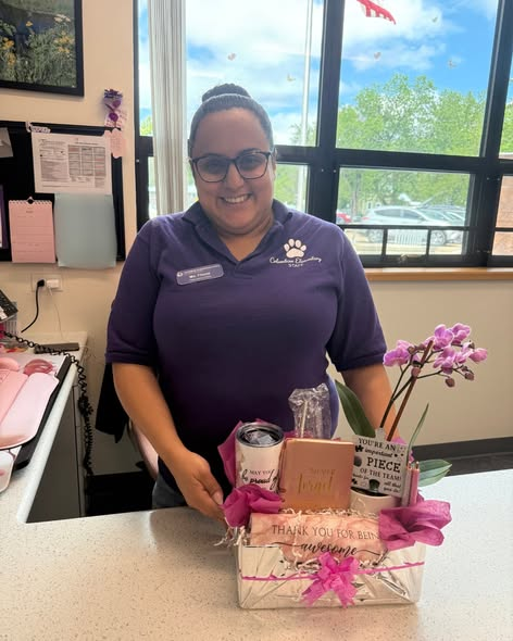 Una mujer sonriente, vestida con una camiseta morada, sostiene una cesta de regalo con flores y una tarjeta, de pie junto al mostrador de recepción.