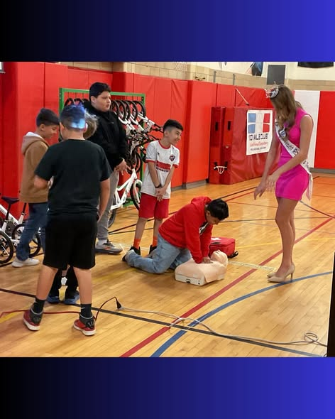 A group of children watches as one boy practices CPR on a dummy, guided by a woman in a pink dress and crown.