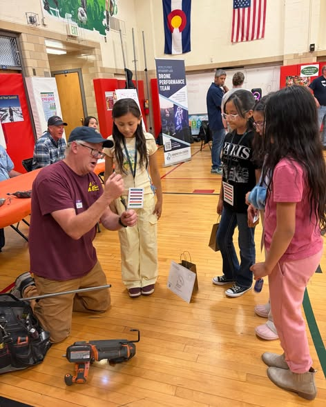 A man demonstrates a tool to three children at a school event, with banners and people in the background.