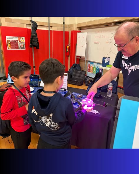 Two children interact with a man at a science fair booth, exploring a glowing device on a table.